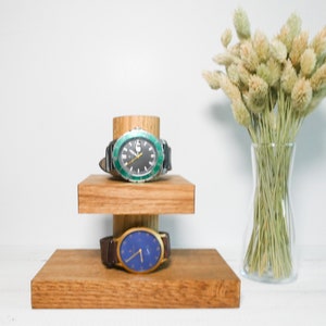 May include: Two watches on a wooden stand with two tiers. The top watch has a black face with a green bezel. The bottom watch has a blue face. The stand is made of light brown wood.