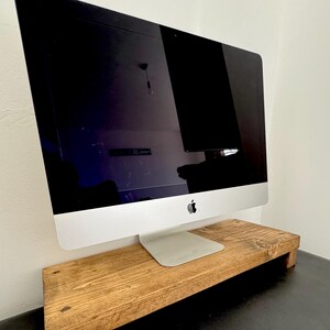 May include: A wooden monitor stand with a natural finish supports a silver iMac computer. The stand is rectangular with a raised platform and short legs. The iMac has a black screen and the Apple logo.