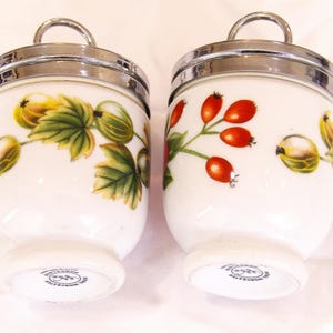 May include: Two white porcelain egg cups with silver-tone rims and handles. One cup features a design of green leaves and yellow-green berries, the other has red berries. The egg cups are from the Worcester brand.