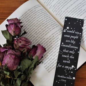Könnte beinhalten: Ein offenes Buch mit einem schwarzen Lesezeichen und getrockneten lila Rosen. Das Lesezeichen hat ein Zitat über Konstellationen und weiße Sterne. Der Text auf dem Lesezeichen lautet: „Ich würde sagen, manche Menschen sind wie Konstellationen, die die Erde nur für eine Saison berühren.“