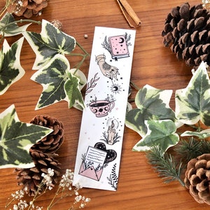 May include: A white bookmark with celestial illustrations, including a book, hand, teacup, candle, and envelope. The bookmark is decorated with a crescent moon and stars. The background includes ivy, pinecones, and sprigs on a wooden surface.