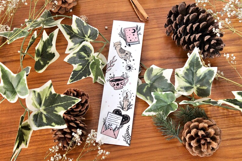 May include: A white bookmark with celestial illustrations, including a book, hand, teacup, candle, and envelope. The bookmark is decorated with a crescent moon and stars. The background includes ivy, pinecones, and sprigs on a wooden surface.