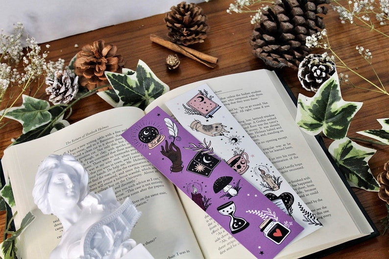 May include: Two decorative bookmarks with celestial and mystical illustrations. One is purple with a crystal ball, hand, and hourglass. The other is white with a book, hand, and teacup. The bookmarks are on an open book with decorative pinecones and greenery.