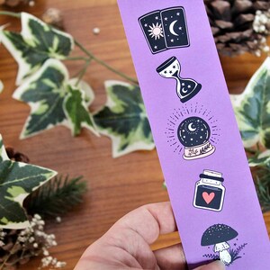 May include: A purple bookmark featuring mystical illustrations. The designs include tarot cards, an hourglass, a crystal ball, a potion bottle with a heart, and a mushroom. The background includes greenery and pinecones.