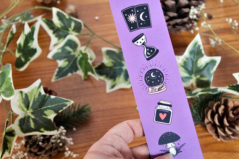 May include: A purple bookmark featuring mystical illustrations. The designs include tarot cards, an hourglass, a crystal ball, a potion bottle with a heart, and a mushroom. The background includes greenery and pinecones.