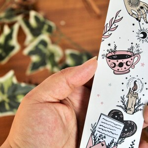 May include: A white bookmark with celestial and botanical illustrations. The design includes a hand, a teacup, a candle, and an envelope. The illustrations are in black and pink, with small stars scattered throughout. The bookmark is held in a hand.