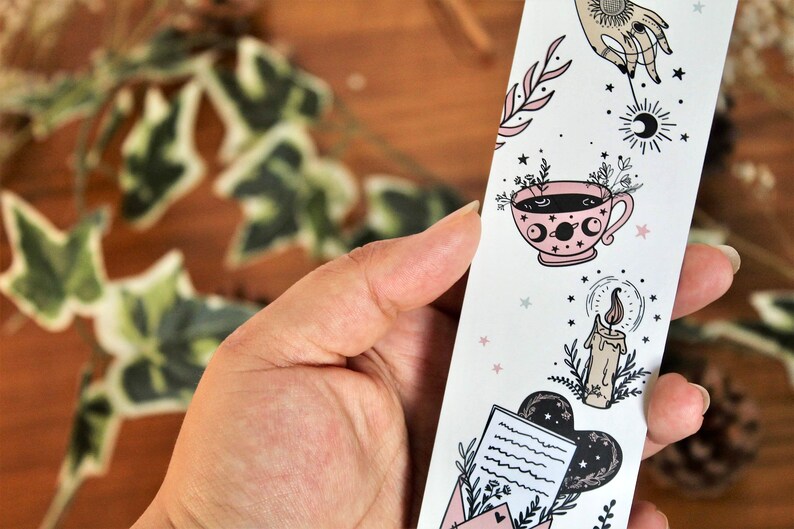 May include: A white bookmark with celestial and botanical illustrations. The design includes a hand, a teacup, a candle, and an envelope. The illustrations are in black and pink, with small stars scattered throughout. The bookmark is held in a hand.