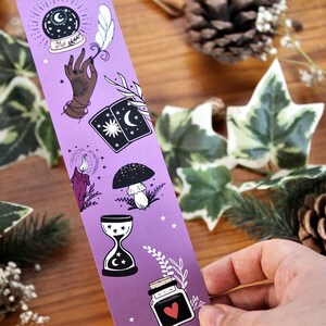 May include: A purple bookmark featuring mystical illustrations. The design includes a crystal ball, hand, tarot cards, candle, mushroom, hourglass, and a potion bottle with a heart. The bookmark is held by a hand, with a wooden background and greenery.