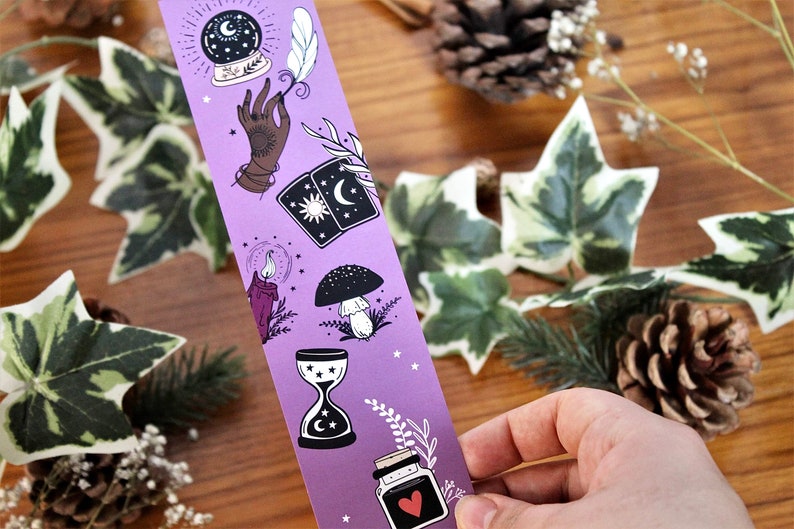 May include: A purple bookmark featuring mystical illustrations. The design includes a crystal ball, hand, tarot cards, candle, mushroom, hourglass, and a potion bottle with a heart. The bookmark is held by a hand, with a wooden background and greenery.