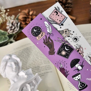 May include: Two decorative bookmarks with celestial and mystical illustrations. One is purple, the other white. The designs include a crystal ball, a hand, a book, a mushroom, a candle, and an hourglass. The bookmarks are placed over an open book.
