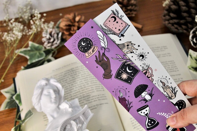 May include: Two decorative bookmarks with celestial and mystical illustrations. One is purple, the other white. The designs include a crystal ball, a hand, a book, a mushroom, a candle, and an hourglass. The bookmarks are placed over an open book.