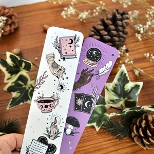 May include: Two decorative bookmarks with celestial and mystical illustrations. One bookmark is white with a pink teacup, a hand with a sun, and a book. The other is purple with a crystal ball, a hand with a feather, and tarot cards.