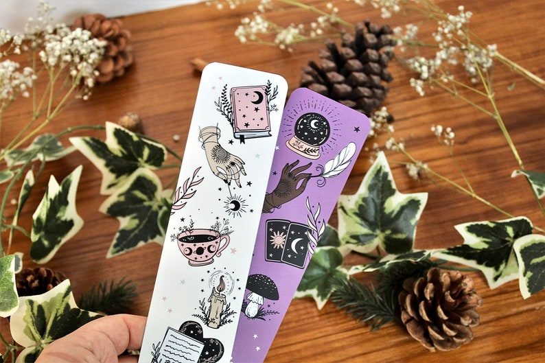 May include: Two decorative bookmarks with celestial and mystical illustrations. One bookmark is white with a pink teacup, a hand with a sun, and a book. The other is purple with a crystal ball, a hand with a feather, and tarot cards.
