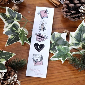 May include: A white bookmark with illustrations of a book, candle, teacup, heart, and letter. The bookmark has the text "Bookishly Divine" at the bottom. The background includes greenery, pinecones, and a wooden surface.