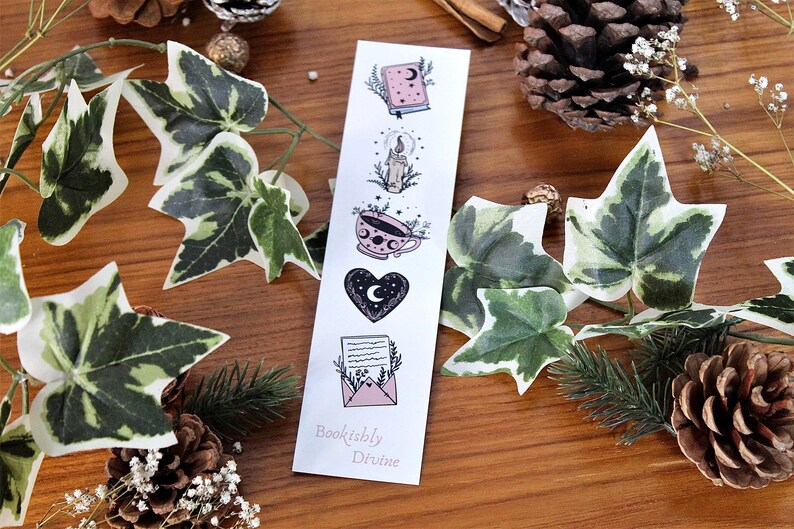 May include: A white bookmark with illustrations of a book, candle, teacup, heart, and letter. The bookmark has the text "Bookishly Divine" at the bottom. The background includes greenery, pinecones, and a wooden surface.