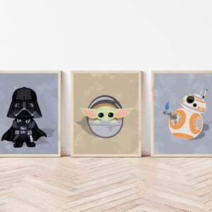 Op de afbeelding: Drie ingelijste prints met Star Wars-personages. Darth Vader in zwart, Baby Yoda in groen en BB-8 in wit en oranje. De prints worden weergegeven op een lichtgrijze achtergrond met houten frames, tegen een lichte houten vloer.