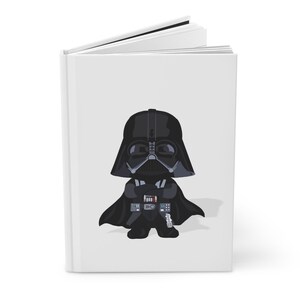Op de afbeelding: Wit hardcover notitieboek met een cartoon illustratie van Darth Vader. Het personage is afgebeeld in zwart met een gedetailleerde helm en cape. Het notitieboek is open en toont blanco witte pagina's.