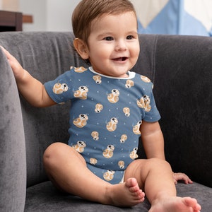 May include: A baby wearing a blue onesie with a white and orange pattern of a robot character. The baby is sitting on a gray couch and smiling.