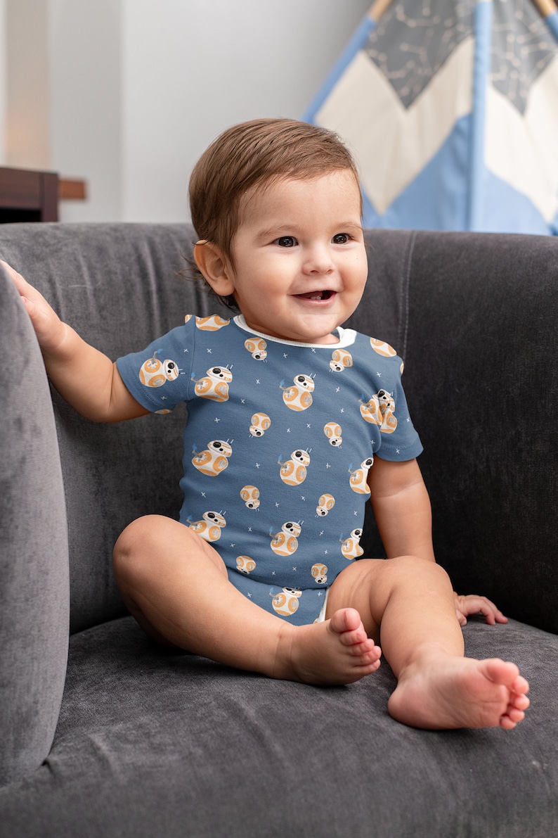 May include: A baby wearing a blue onesie with a white and orange pattern of a robot character. The baby is sitting on a gray couch and smiling.