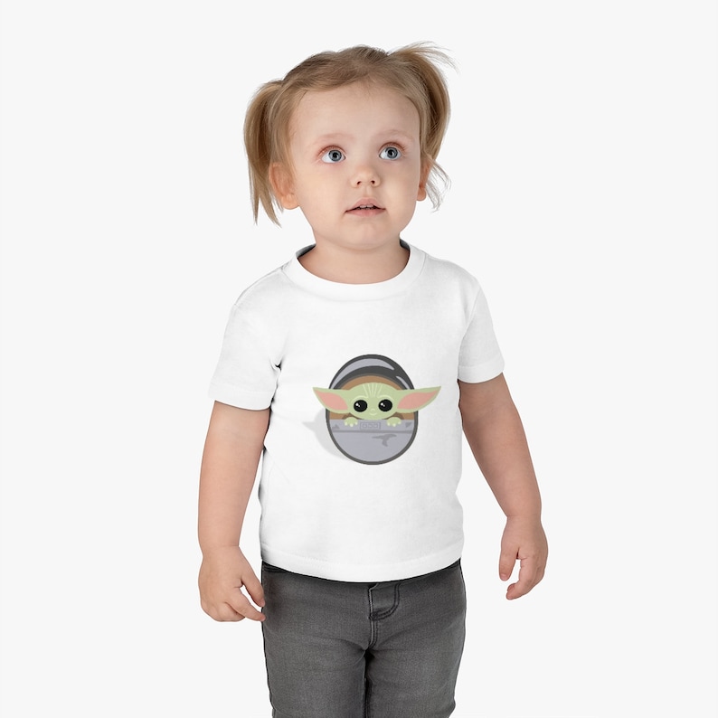 Op de afbeelding: Wit T-shirt met een cartoonfiguur met grote oren en ogen, die uit een grijs ovaal komt. Het personage is groen en beige. Het kind draagt een grijze broek.