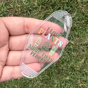 May include: Clear acrylic keychain with the text "I LOVE PLANTS MORE THAN PEOPLE" in colorful lettering. The keychain features illustrations of various plants. A silver key ring is attached to the top.
