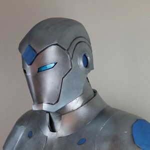 May include: A silver and blue superhero costume, featuring a helmet with a diamond-shaped blue accent and glowing blue eyes. The chest and shoulders have blue circular and angular details. The overall design is metallic and futuristic.
