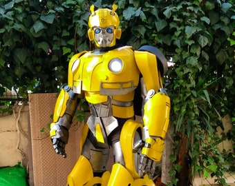 Puede incluir: Un disfraz de cuerpo completo de robot amarillo y gris, con un diseño detallado. El robot tiene un casco con antenas, un pecho con dos luces circulares y extremidades grandes y articuladas. El disfraz está de pie sobre una superficie de piedra.