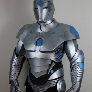 May include: A silver and blue superhero costume. The costume features a helmet, chest armor, and arm and leg coverings. Blue accents are visible on the shoulders, arms, and chest. The suit is made of a metallic material.