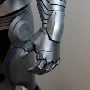 May include: Close-up of a silver and blue armored costume. The image focuses on the articulated hand and forearm, showcasing the metallic texture and segmented design. The costume appears to be part of a larger ensemble.