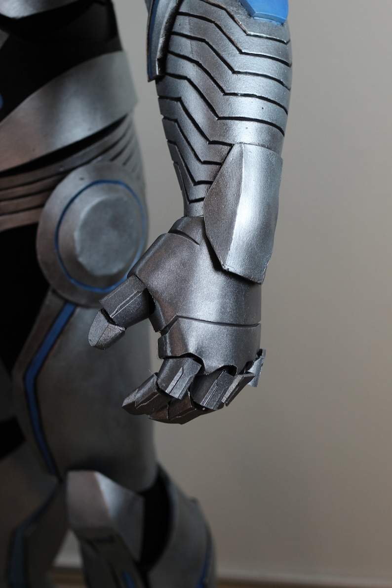 May include: Close-up of a silver and blue armored costume. The image focuses on the articulated hand and forearm, showcasing the metallic texture and segmented design. The costume appears to be part of a larger ensemble.