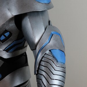 May include: Close-up of a silver and blue armored costume. The shoulder and arm sections are visible, showcasing a metallic finish with blue accents. The design features layered plates and geometric shapes.