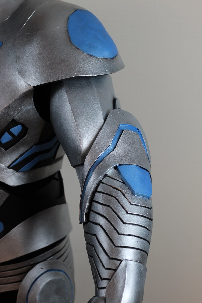 May include: Close-up of a silver and blue armored costume. The shoulder and arm sections are visible, showcasing a metallic finish with blue accents. The design features layered plates and geometric shapes.