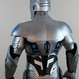 May include: A silver and blue futuristic costume, viewed from the back. The helmet and armor feature intricate paneling and detailing. Blue accents highlight the shoulders, back, and arms. The overall design suggests a science fiction or cosplay theme.
