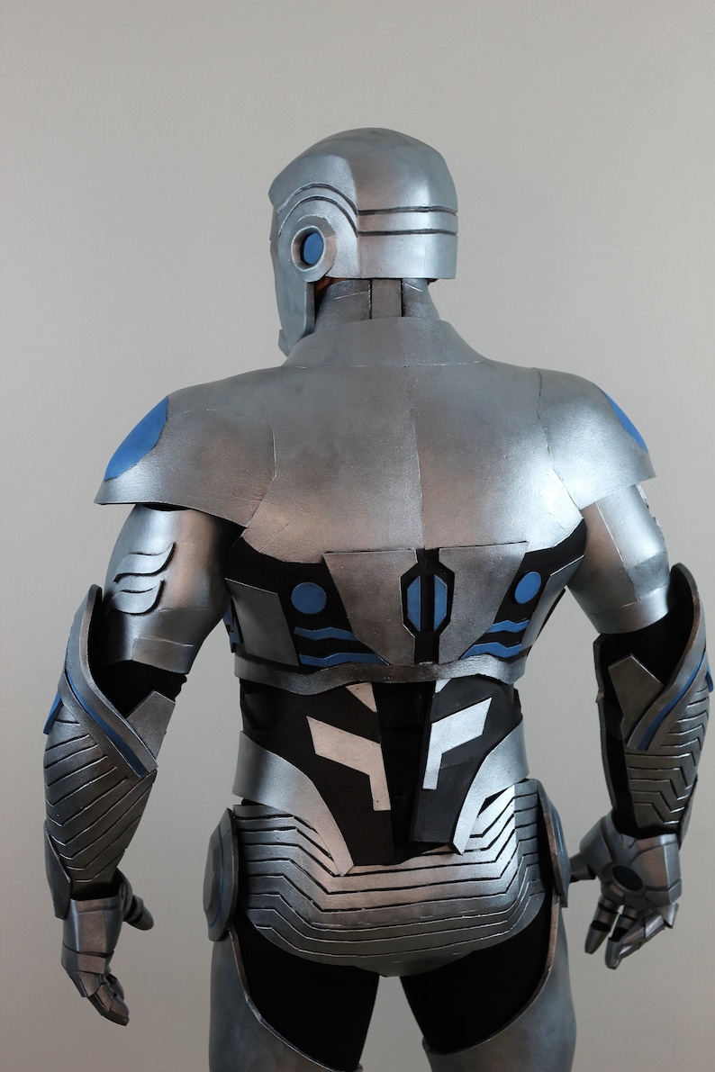 May include: A silver and blue futuristic costume, viewed from the back. The helmet and armor feature intricate paneling and detailing. Blue accents highlight the shoulders, back, and arms. The overall design suggests a science fiction or cosplay theme.