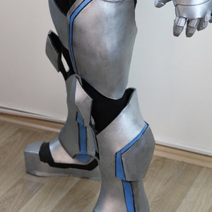 May include: Silver and black robotic leg armor with blue accents. The armor features articulated joints, protective plating, and a futuristic design. The boots have a blocky shape, and the overall aesthetic is reminiscent of a superhero or science fiction costume.