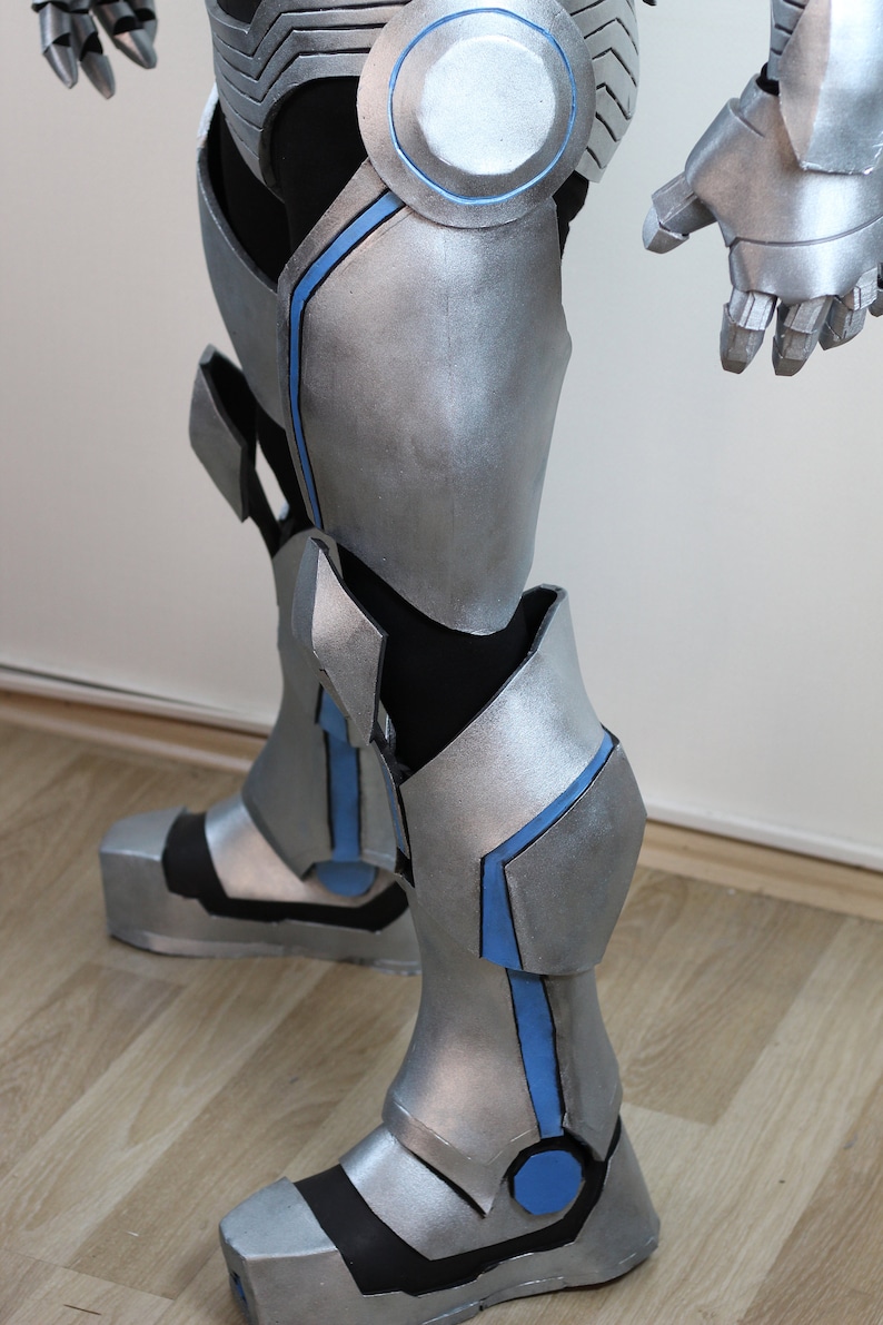 May include: Silver and black robotic leg armor with blue accents. The armor features articulated joints, protective plating, and a futuristic design. The boots have a blocky shape, and the overall aesthetic is reminiscent of a superhero or science fiction costume.