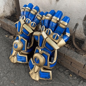 May include: A pair of blue and gold gauntlets, likely for cosplay or costume use. The gauntlets feature a detailed design with gold accents on the knuckles, wrist, and other areas. The blue sections have a metallic sheen.