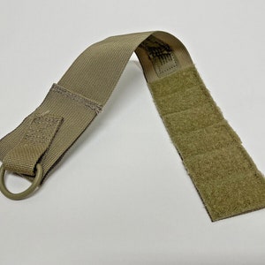 Puede incluir: Una correa táctica de color marrón claro con un anillo en D y un cierre de gancho y bucle. La correa está hecha de nylon y tiene una superficie texturizada.