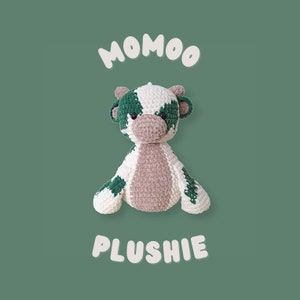 May include: A green and white crocheted cow plush toy with the text "MOMOO PLUSHIE" on a green background.