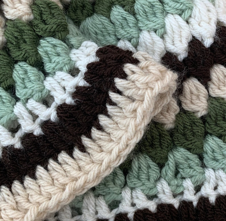 May include: Close-up of a crocheted blanket with brown, white, and green stripes. The blanket is made with a textured stitch that creates a raised pattern.