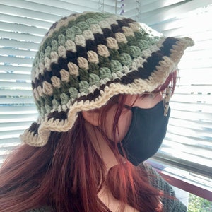 May include: A crocheted bucket hat with brown, green, and white stripes. The hat has a scalloped brim and is worn by a person with long, red hair.