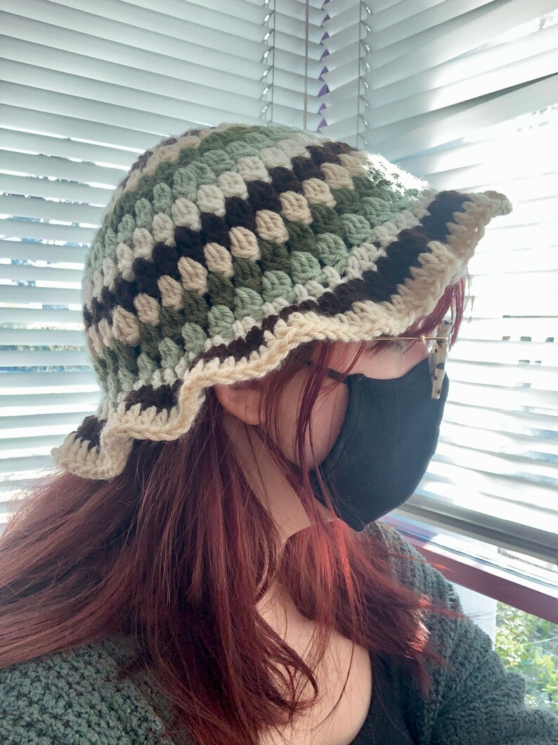 May include: A crocheted bucket hat with brown, green, and white stripes. The hat has a scalloped brim and is worn by a person with long, red hair.