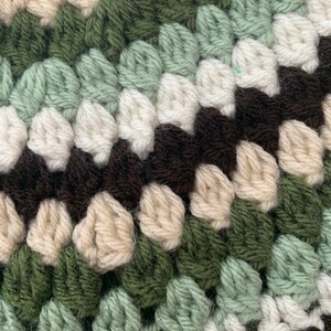 May include: Close-up of a crocheted fabric with a repeating pattern of brown, cream, green, and light green yarn.