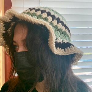 May include: A person wearing a black face mask and a crocheted bucket hat with green, white, and black stripes. The hat has a wide brim and a scalloped edge.