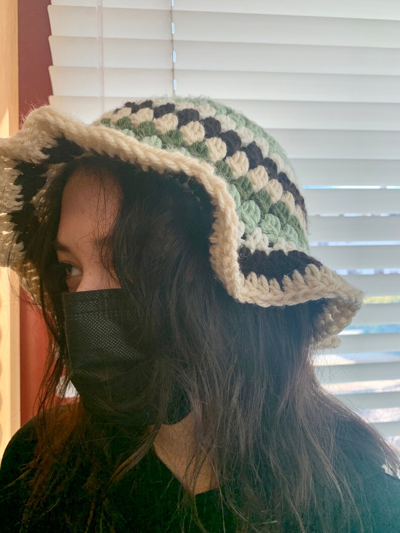 May include: A person wearing a black face mask and a crocheted bucket hat with green, white, and black stripes. The hat has a wide brim and a scalloped edge.