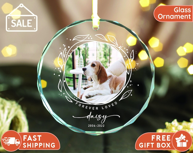 May include: A clear glass ornament with a white floral design and a photo of a dog. The ornament has the text "Forever Loved Daisy 2004-2022" inscribed on it.