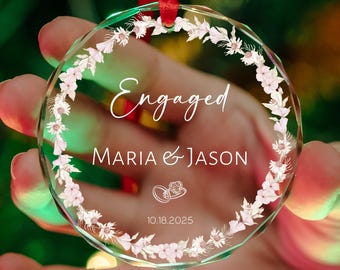 May include: A clear, round glass ornament with the word "Engaged" in elegant script. The names "MARIA & JASON" are below, along with a date and ring graphic. The ornament is edged with a floral design and has a red ribbon for hanging.