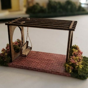 May include: A miniature wooden swing set with a brown wooden swing hanging from a pergola. The swing set is surrounded by green and pink miniature plants and sits on a brick patio.