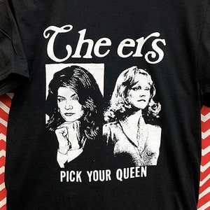 May include: Black t-shirt with a white graphic of two women from the TV show "Cheers" with the text "The ers" and "Pick Your Queen".