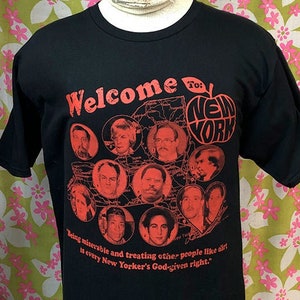 May include: Black t-shirt with a red graphic print of an apple with the words "Welcome to: New York" and a map of New York State. The graphic also includes a collage of faces in red circles. The text below the graphic reads "Being miserable and treating other people like air is every New Yorker's God-given right."